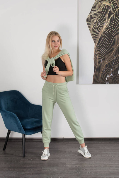 Stylish beautiful young blond woman in a light green tracksuit poses near a white wall in the room. Attractive girl model posing near blue chair. - 写真・画像