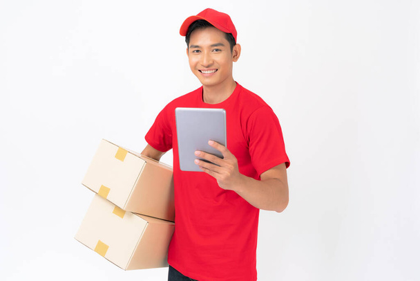 Usmívající se doručovatel zaměstnanec v červené čepici prázdné tričko uniforma stojící s balíčkem poštovní schránka izolované na bílém pozadí - Fotografie, Obrázek