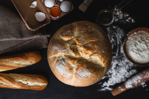 小麦粉、卵、キッチン用品のボウルと黒石のテーブルの上に新鮮な焼きたてのパンを粉砕します。トップビュー、フラットレイアウト - 写真・画像