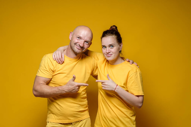 Lächelnder Mann und Frau in schlichter, heller Kleidung umarmen sich und zeigen mit dem Zeigefinger auf einen gelben Hintergrund. - Foto, Bild
