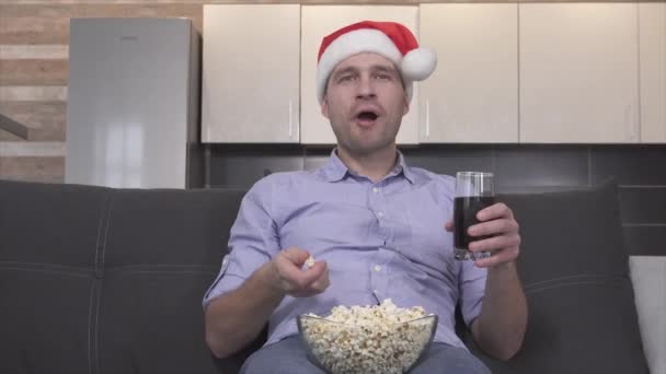 Emotionaalinen juhlava nuori mies jouluhatussa syö popcornia ja juo limsaa television, elokuvan tai elokuvan aikana. Sir yksin sohvalla ja vaihtaa kanavaa kaukosäätimellä. Emotionaalinen onnellinen kaveri. - Materiaali, video