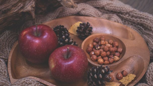 Autumn still life with apples and nuts. Autumn background with apples on a warm knitted scarf, a wooden plate, autumn leaves, hazelnuts and cones. - Photo, image