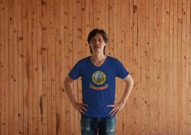 Man wearing Idaho flag color shirt and standing with akimbo on the wooden wall background. State seal of Idaho on a field of blue.  - Photo, Image