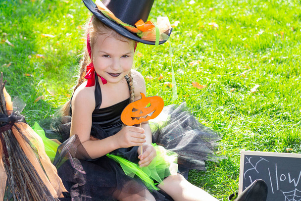 girl in a witch costume for the Halloween holiday. Placard with the inscription: Halloween. Girl sitting on the grass with pumkin - Foto, imagen
