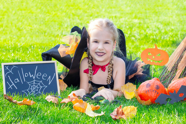 girl in a witch costume for the Halloween holiday. Placard with the inscription: Halloween. Happy girl lies on the grass - Fotoğraf, Görsel