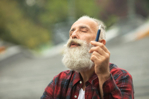 Senior man with beard breathe out smoke. Smoking electronic cigarette. Stress relief concept. Smoking device. Man long beard relaxed with smoking habit. Bearded man vaping. IQOS. - Фото, изображение