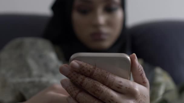 Mujer musulmana navegando en su teléfono inteligente en interiores sentada. Enfoque superficial, ángulo bajo, bloqueado - Metraje, vídeo