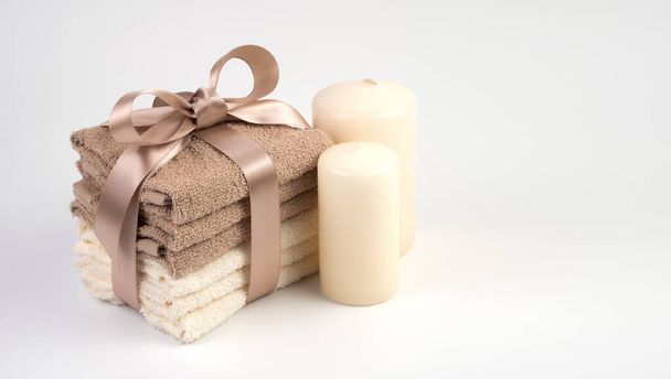 Подарочный набор с полотенцами Терри, завязанными атласной лентой и свечами на светлом фоне. Вид сбоку. - Фото, изображение