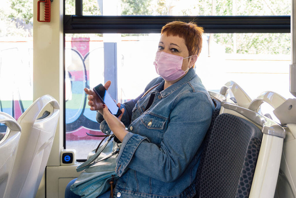 Redhead middle-aged woman with mandatory mask inside public spaces for transportation like bus using smartphone - Foto, Imagem