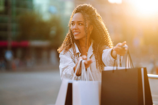 Elegant African American woman holds out shopping bags. Young woman with packages after shopping. Purchases, black friday, discounts, sale concept. - Photo, image
