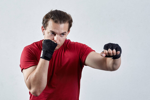 Young man boxes, making jab with left hand. Black bandages, red T-shirt, determined face expression, strong emotions. Concept of blow to kill or hit, copy space on fist and on light background. - Φωτογραφία, εικόνα