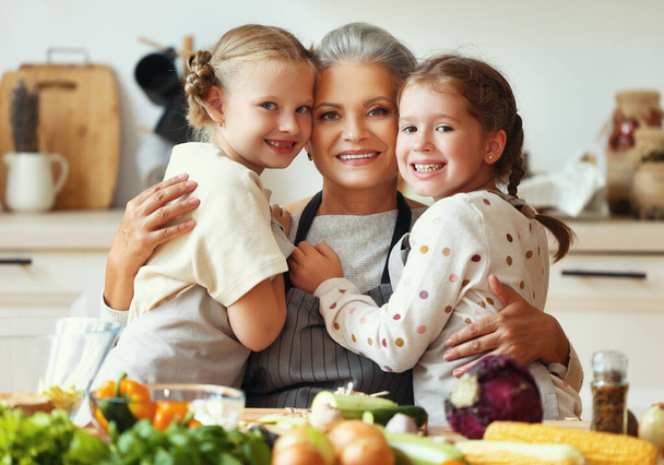 Gelukkig gezin: schattige kleine meisjes in schorten knuffels gelukkig grootmoeder in voorbereiding van gezonde vegetarische schotel met verse ingrediënten in huis kitche - Foto, afbeelding