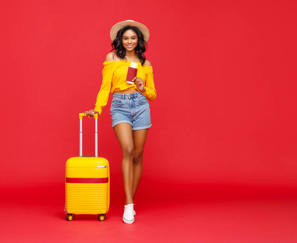 Mulher étnica de corpo inteiro na moda com passaporte e bilhetes sorrindo para câmera e apoiando-se na bagagem durante as férias de verão contra o backdro vermelho - Foto, Imagem