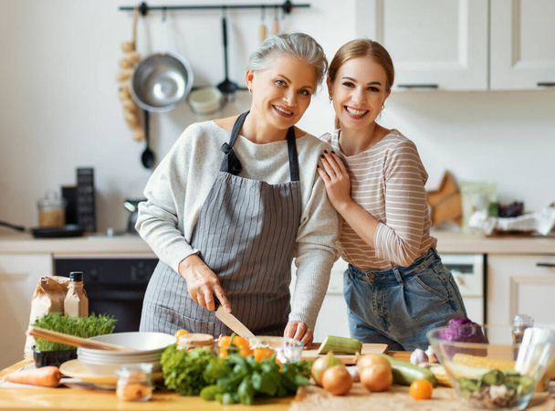 幸せな家族明るい若い女性は、家庭のキッチンで新鮮な野菜と健康的な料理を準備しながら、成熟した母親を受け入れ - 写真・画像