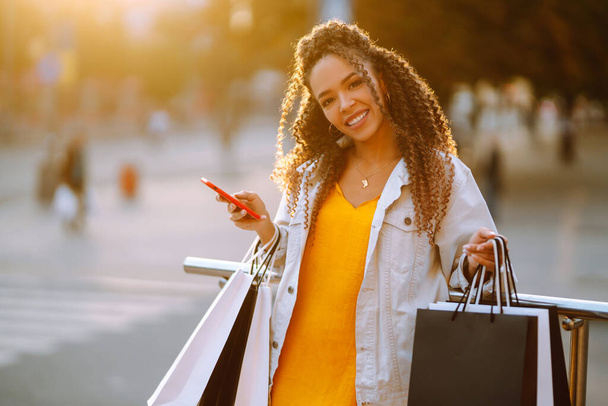 African american woman shopping with a phone in european city street at sunset. Young woman standing along the road holding shopping bags and using mobile phone. Purchases, black friday, discounts. - Foto, immagini