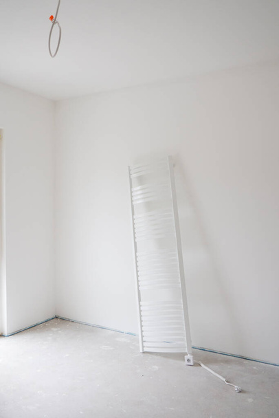 Verticale opname van lege witte kamer in huis tijdens renovatie.Unconnected grote verticale elektrische verwarmde handdoekenrek van klassieke laddervorm staat tegen de tegenovergestelde muur.Badkamer renovatie.Kopieer ruimte. - Foto, afbeelding