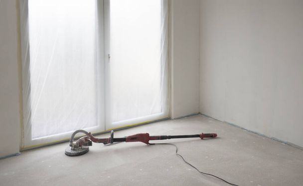 Schmutziger und staubiger roter Schleifer für Wände und Decken liegt auf dem Boden im leeren Raum vor weißen anstrichfertigen Wänden und deckenhohen französischen Fenstern.. - Foto, Bild