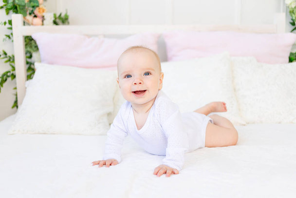 Ευτυχισμένο κοριτσάκι 6 μηνών, με λευκή φόρμα ξαπλωμένη μπρούμυτα σε λευκό κρεβάτι στο σπίτι. - Φωτογραφία, εικόνα