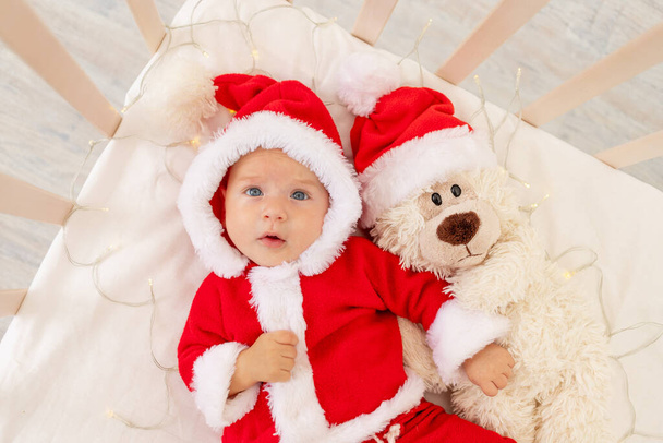 Kerstfoto van een baby in een kerstman kostuum liggend in een wieg thuis met een speelgoed in een kerstman hoed, bovenaanzicht, gelukkig nieuwjaar. - Foto, afbeelding