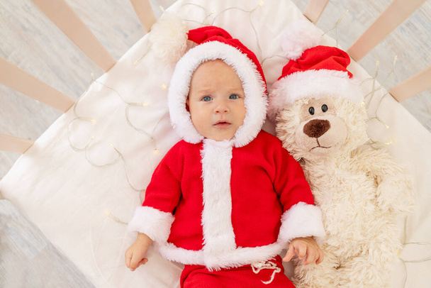 Weihnachtsfoto eines Babys im Weihnachtsmannkostüm, das zu Hause in einer Krippe liegt, mit einem Spielzeug im Nikolausmütze, Draufsicht, frohes neues Jahr. - Foto, Bild