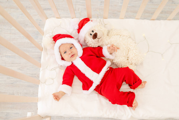 Christmas photo of a baby in a Santa costume lying in a crib at home with a toy in a Santa Claus hat, top view, happy new year. - Foto, Imagem