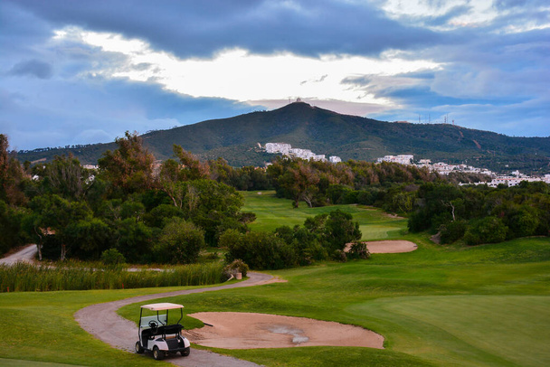 Καρότσι γκολφ σε γήπεδο γκολφ. Πράσινο πεδίο και συννεφιασμένος μπλε ουρανός. Ανοιξιάτικο τοπίο με γρασίδι και δέντρα - Φωτογραφία, εικόνα