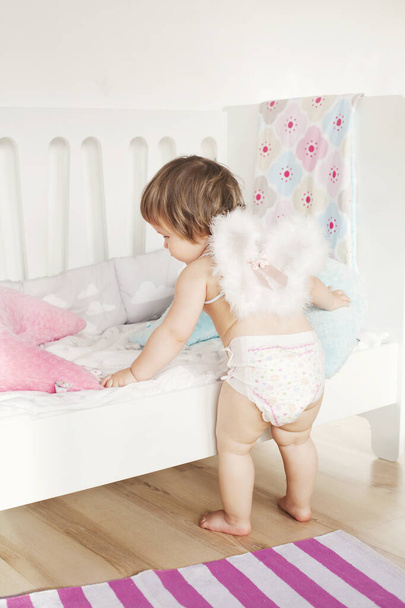 Todler κοριτσάκι σε πάνα και φτερά αγγέλου παίζει κοντά στο κρεβάτι. Εσωτερικός σχεδιασμός παιδικού δωματίου σε ανοιχτόχρωμα χρώματα - Φωτογραφία, εικόνα