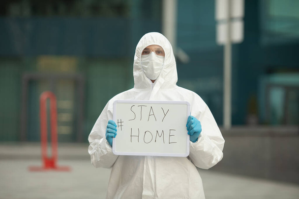 Doctor in protective costume and face medical mask standing on street and holding board with text to stay at home during pandemic. Covid-19. Coronavirus. - Zdjęcie, obraz