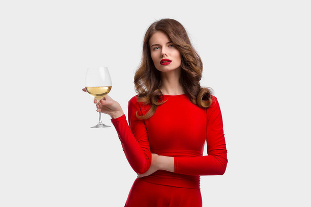Woman with makeup, hairstyle waering red dress posing with glass of vine over white background, isolate - Foto, Imagen