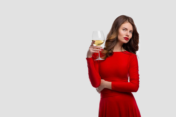 Woman with makeup, hairstyle waering red dress posing with glass of vine over white background, isolate - Zdjęcie, obraz