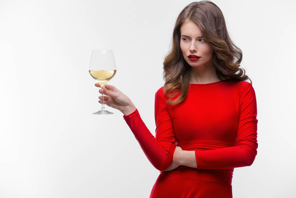 Woman with makeup, hairstyle waering red dress posing with glass of vine over white background, isolate - Photo, Image