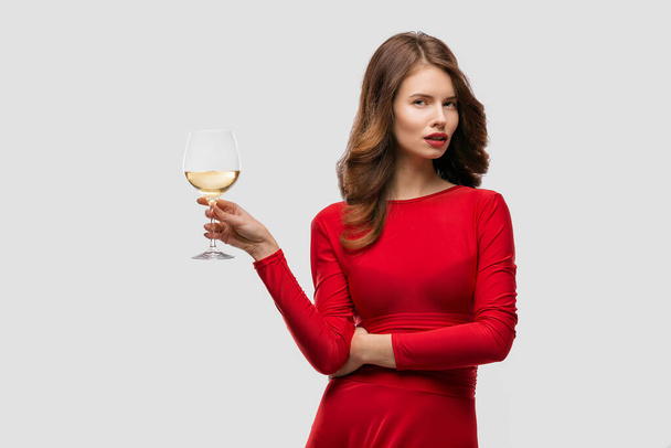 Woman with makeup, hairstyle waering red dress posing with glass of vine over white background, isolate - Valokuva, kuva