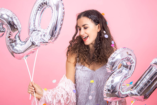 Beautiful young brunette girl with curly hair and festive clothes posing on a pink studio background with confetti and holding in her hand silver balloons from the numbers 2021 close up - Фото, изображение