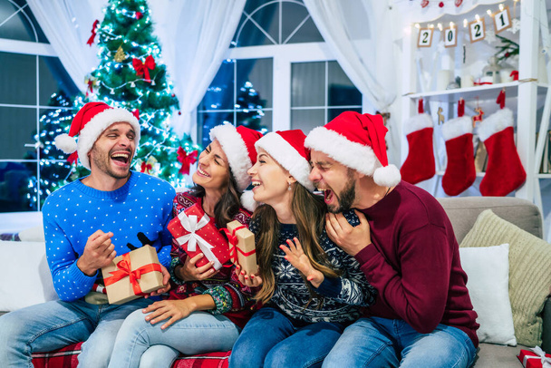 Heyecanlı ve gülen genç arkadaşlar Noel tatilini dekore edilmiş bir evde, arka planda büyük parlak bir Noel ağacıyla kutluyorlar. - Fotoğraf, Görsel