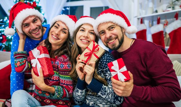 Group of beautiful best friends in Santa hats with gifts in hands are posing and having fun on Christmas tree background at home party - Foto, Bild
