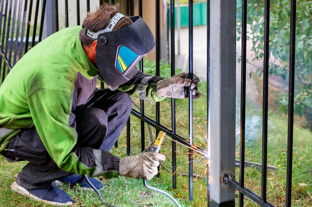 Ένας συγκολλητής που φοράει προστατευτικά ρούχα, κράνος και γάντια χρησιμοποιώντας ένα ηλεκτρόδιο συγκολλά ένα μεταλλικό φράχτη γύρω από την περιοχή εργασίας, στήνοντας ένα φράχτη, επιλεκτική εστίαση, αντιγραφή χώρου. - Φωτογραφία, εικόνα