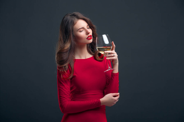Woman with bright makeup, hairstyle wearing red dress posing with glass of vine over dark background, isolate - Zdjęcie, obraz