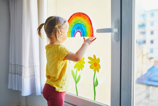 Adorable toddler girl attaching drawing of rainbow to window glass as sign of hope. Creative games for kids staying at home. Self isolation and quarantine concept - Φωτογραφία, εικόνα