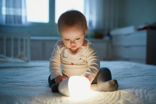Adorable baby girl playing with bedside lamp in nursery. Happy kid sitting on bed with nightlight. Little child at home in the evening before sleep - Fotoğraf, Görsel