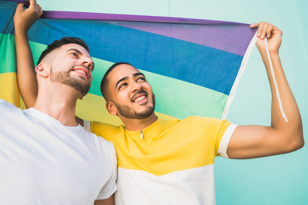 Portrait of young gay couple embracing and showing their love with rainbow flag in the street. LGBT and love concept - Photo, image