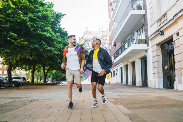 Portrait of young gay couple holding their hands and running together with rainbow flag in the street. LGBT and love concept - 写真・画像