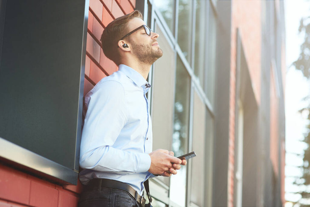 Favourite song. Side view of a young happy businessman listening to music with wireless earphones and mobile phone while standing outdoors - Photo, Image