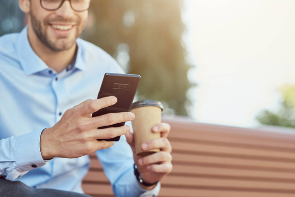 Online chattailu. Sivukuva onnellinen liikemies älypuhelimella ja kahvinjuonti samalla rentoutuen penkillä ulkona - Valokuva, kuva