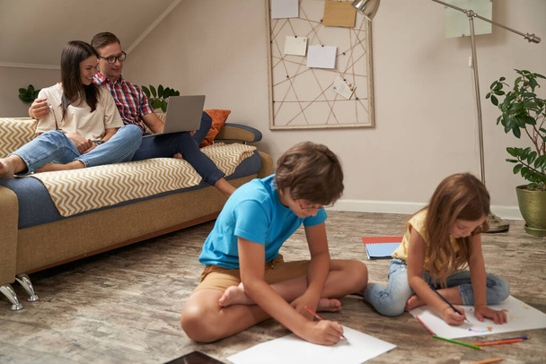 Pieni poika ja tyttö istuu puulattialla ja piirtää värikkäitä kyniä, kun heidän vanhempansa, onnellinen äiti ja isä rentoutuvat sohvalla ja käyttävät kannettavaa tietokonetta. - Valokuva, kuva