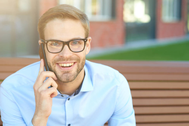 Portrait of a young happy businessman talking by phone and and smiling at camera while sitting on the bench outdoors - 写真・画像