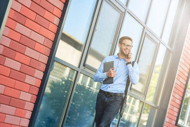 Молодий щасливий кавказький бізнесмен у блакитній сорочці та окулярах тримає ноутбук, розмовляє по телефону та посміхається під час прогулянки на відкритому повітрі
 - Фото, зображення