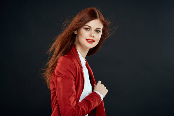 Pretty woman with long hair red jacket makeup lips charm dark background - Фото, изображение