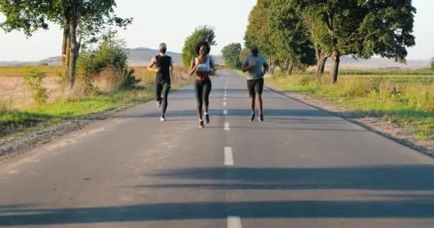 Mladí muži a ženy běhají v létě po venkovské silnici. Afroameričanka štíhlá sportovní žena a multietničtí muži za běháním venku. Běžci v maskách během pandemie. - Záběry, video