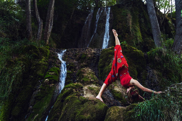 Vista frontal retrato de mujer caucásica adulta de 30 años sentada sobre roca en la naturaleza frente a cascada en vestido rojo estirándose en pose de yoga asana - Espiritualidad concepto de paz interior y relajación - Foto, imagen
