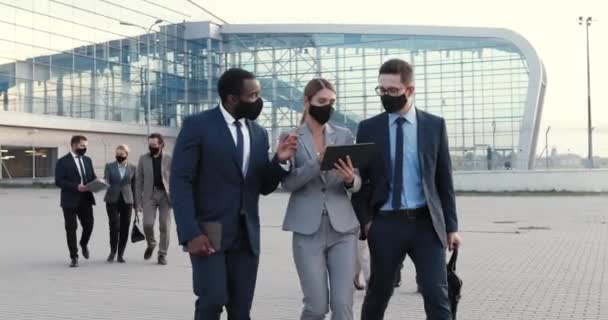 Caucasian businesswoman and two mixed-races businessmen in masks walking outdoor at business center and talking with tablet device in hands. Multi ethnic work partners discussing project and finance. - Footage, Video